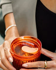 A close-up of a person holding an amber-coloured reusable coffee cup with a same coloured lid and an integrated seal. The person is wearing a gray blazer over a black top, with a pearl bracelet and rings visible.