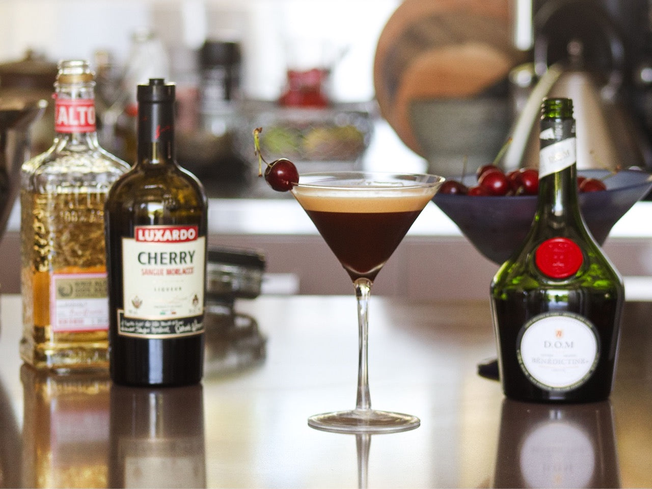 A coffee based cocktail sitting on a table next to bottles of liqueur and a bowl of cherries