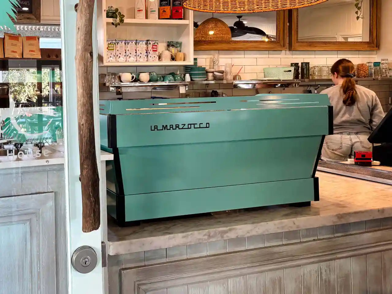 Aqua La Marzocco coffee machine in a cafe setting with a barista in the background.