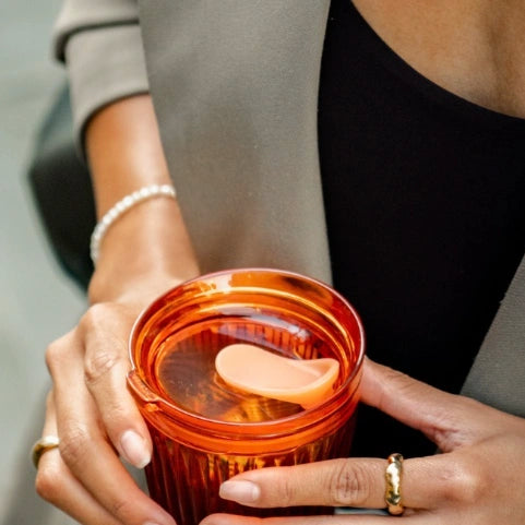 A close-up of a person holding an amber-coloured reusable coffee cup with a same coloured lid and an integrated seal. The person is wearing a gray blazer over a black top, with a pearl bracelet and rings visible.