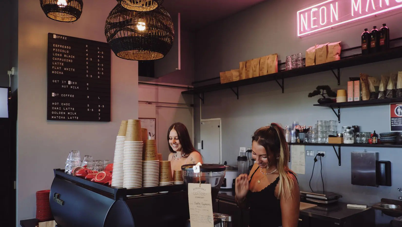 Two women working behind a counter in a coffee shop with neon signs and shelves.