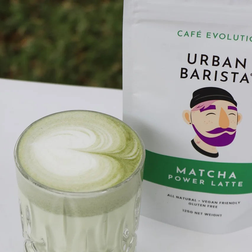 Matcha power latte in a glass with Urban Barista packaging on a white surface.