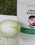Matcha power latte in a glass with Urban Barista packaging on a white surface.