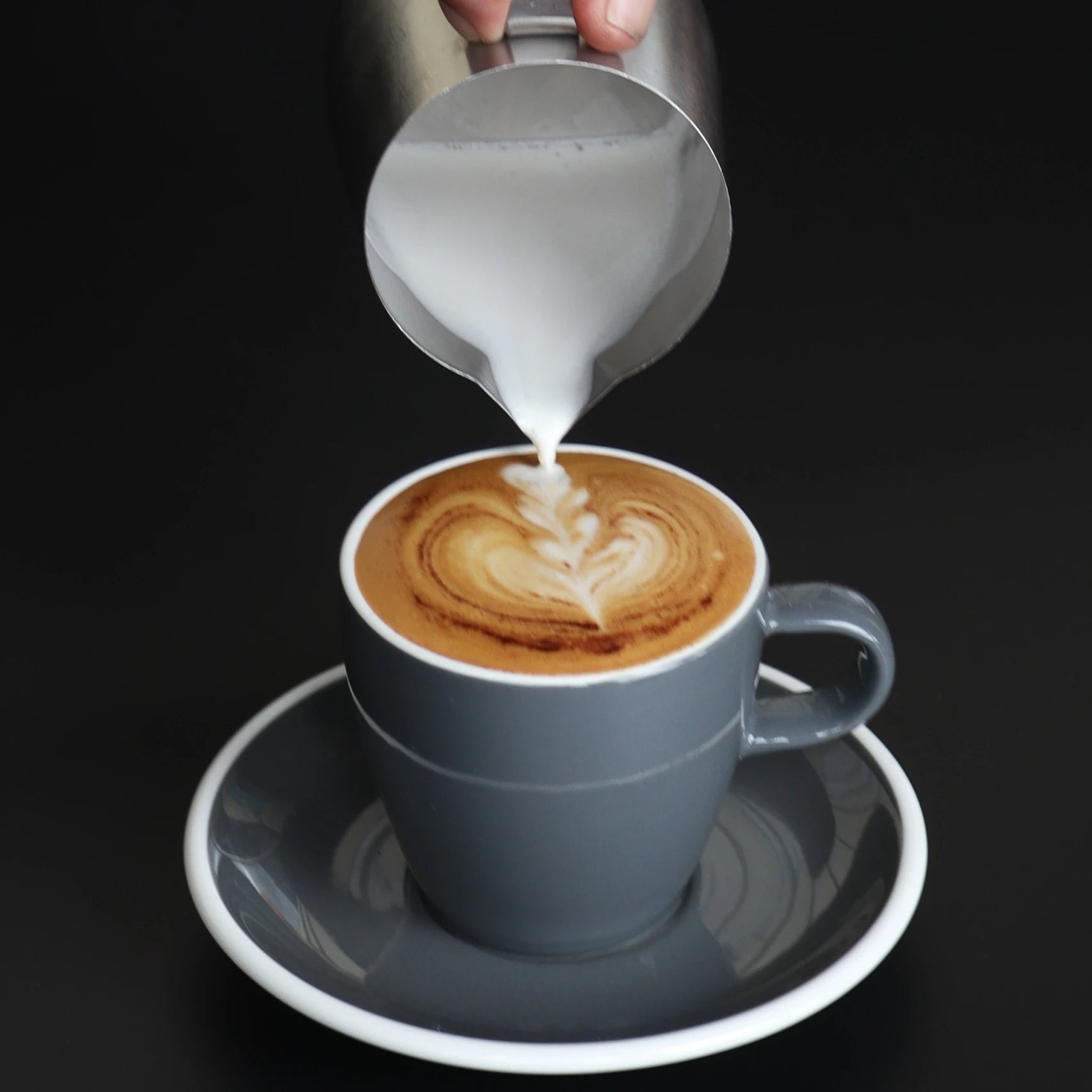 Cappuccino being prepared with heart-shaped design, being poured into a gray cup on a black background.
