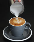 Cappuccino being prepared with heart-shaped design, being poured into a gray cup on a black background.